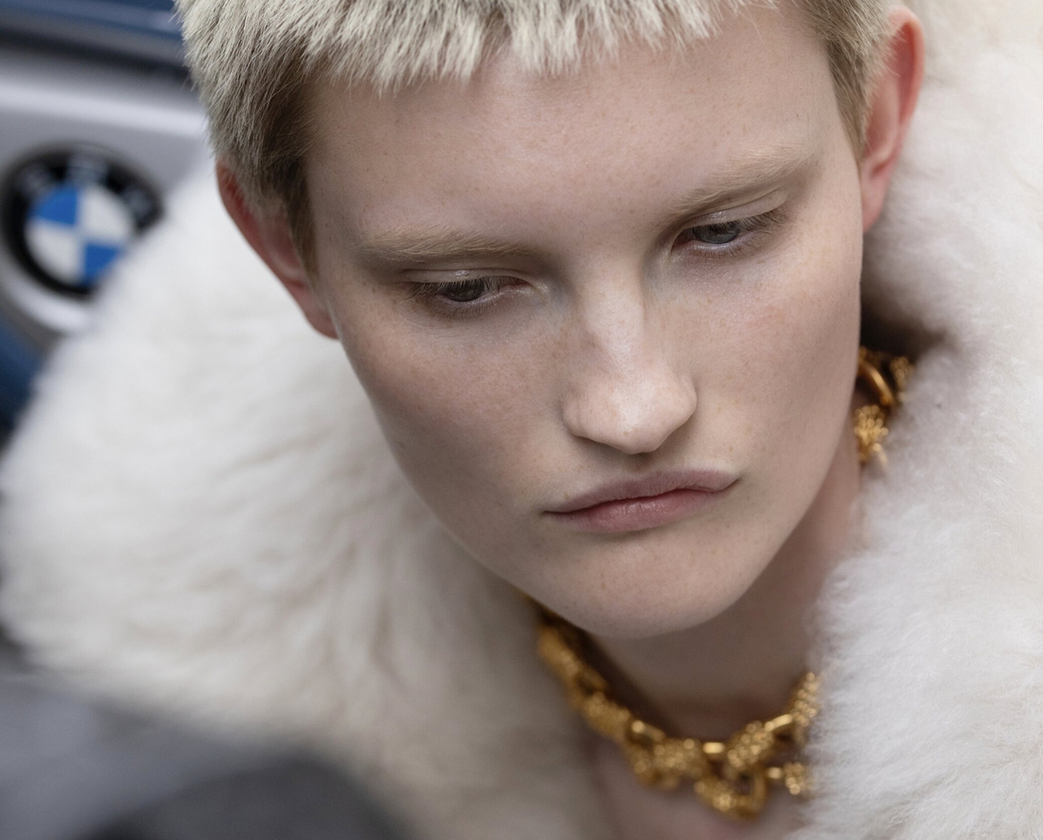 Close-up portrait of a model with cropped platinum hair, freckled skin, a chunky gold chain necklace and white fur collar