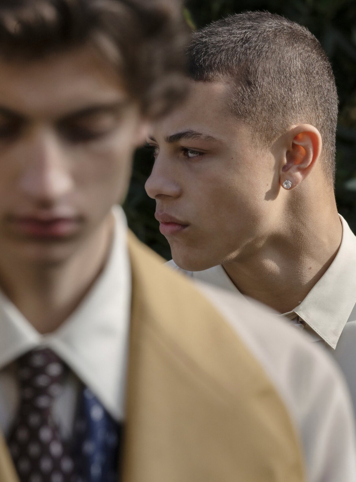 Close profile portrait of a model in a tan jacket beside an out-of-focus model in a tie